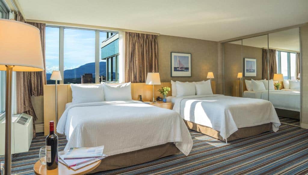 Quarto no Blue Horizon Hotel com uma cama de casal e uma de solteiro, do lado esquerdo há um armário em que as portas têm espelhos, o carpete é listrado em azul, branco e marrom, uma cabeceira de madeira interliga as camas e, por trás delas, há uma janela com cortinas dando vista para montanhas.