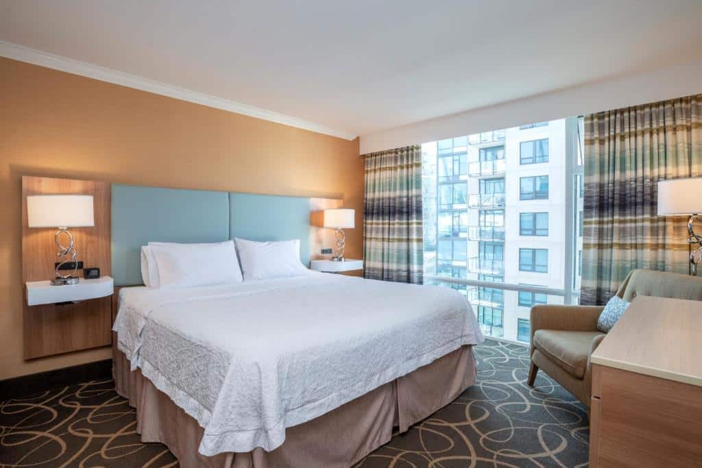 Quarto do Hampton Inn & Suites, by Hilton - Vancouver Downtown com uma janela com cortinas do lado direito, com uma poltrona e um abajur de chão ao lado, na parte esquerda do quarto, está uma cama de casal com uma cabeceira que inclui duas mesinhas com abajures sob elas.