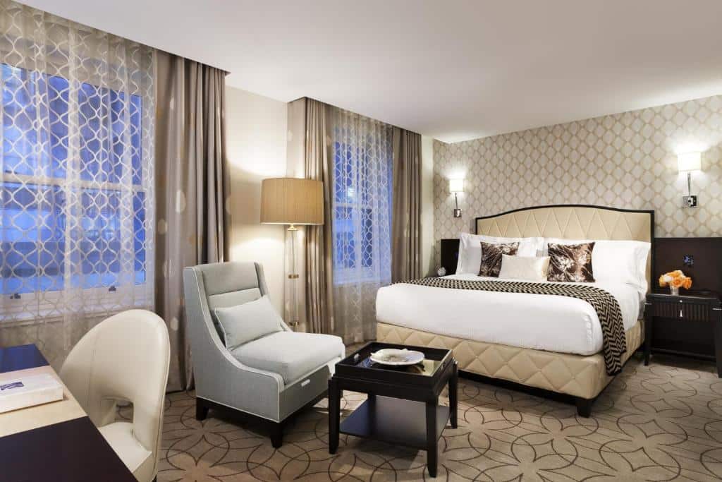 Quarto do Rosewood Hotel Georgia, há duas janelas com cortinas, uma cama de casal com almofadas, na frente da cama tem uma poltrona cinza, o chão é de carpete bege com desenhos minimalistas em marrom, o quarto é inteiro decorado em tons de bege e tons de madeira, para representar hotéis em Vancouver.