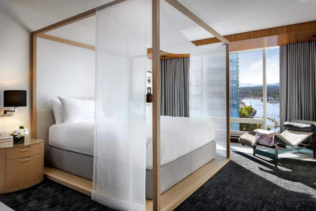 Quarto no Fairmont Pacific Rim com uma cama de casal estilo bangalô, do lado direito há uma ampla janela com vista para a baía, e do lado dessa janela há uma espreguiçadeira de couro. o chão é um carpete cinza escuro.
