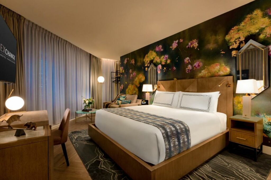 Quarto do EXchange Hotel Vancouver decoração com um papel de parede com rosas desenhadas atrás da cama de casal, duas mesinhas de cabeceira de madeira com abajures em cima delas, o chão é de madeira, do lado esquerdo tem uma ampla janela com uma poltrona, um abajur de chão e uma pequena mesinha de centro, de frente com a cama, há uma mesa de escritório com uma cadeira, e uma televisão presa na parede sob a mesa, para representar hotéis em Vancouver.