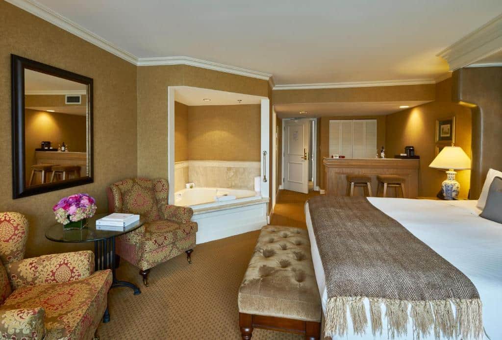 Quarto amplo do Wedgewood Hotel & Spa - Relais & Chateaux, do lado esquerdo há uma banheira de hidromassagem, ao lado, uma pequena mesinha com duas poltronas acolchoadas e um espelho, do lado direito, há um armário embutido e um pequeno balcão com dois bancos altos para se sentar, além da cama de casal com uma mesinha de cabeceira em que há um abajur sob ela, para representar hotéis em Vancouver.