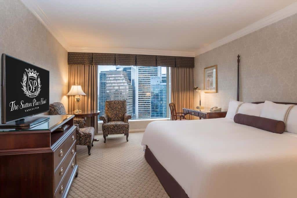 Quarto no The Sutton Place Hotel Vancouver com uma janela ampla com cortinas que dá vista para a cidade, perto da janela há uma pequena mesinha de madeira com um abajur em cima, ao redor dela tem duas poltronas estofadas com flores, o chão é de carpete, há uma cômoda de madeira do lado esquerdo com uma televisão em cima e, do lado esquerdo, há uma cama de casal, próximo dela tem uma mesinha de escritório com uma cadeira.