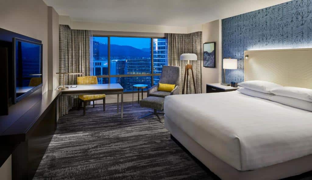 Quarto no Hyatt Regency Vancouver, o local é amplo, há uma janela grande com cortinas e vista para a cidade, uma mesa de escritório com uma cadeira e uma poltrona cinza, o chão é coberto por um carpete cinza, há uma televisão de frente para a cama de casal que tem uma cabeceira branca com iluminação indireta presa nela, a parede atrás da cama tem papel de parede azul com detalhes preto.