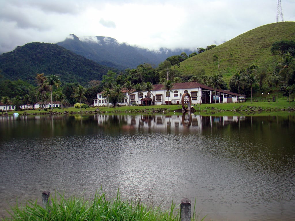 Lago com uma margem gramada, uma casa branca e uma roda d'água, cercado por colinas e árvores sob um céu nublado.