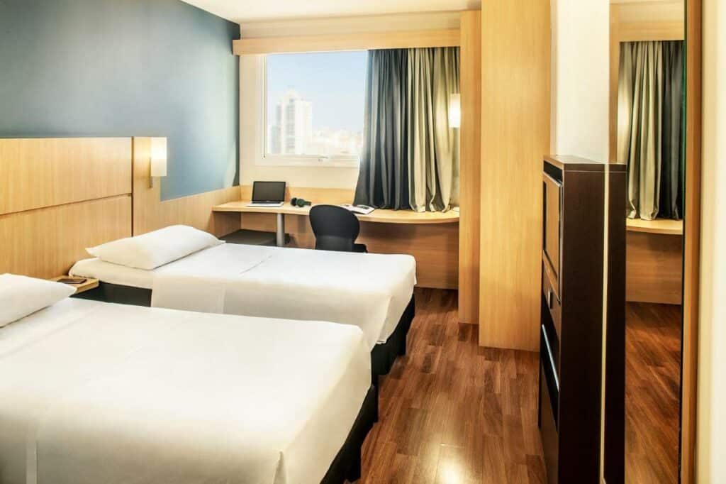 Quarto de hotel com duas camas de solteiro com lençóis brancos, janela com cortina e mesa abaixo da janela com abajour, notebook e cadeira. 