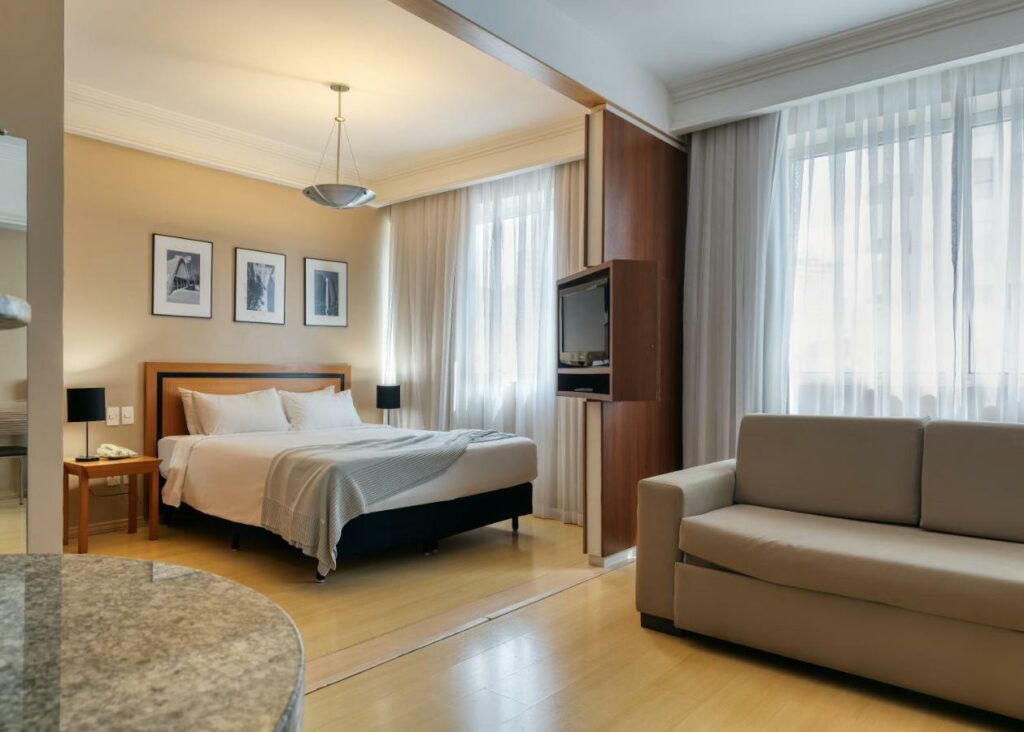Quarto do hotel Mercure Belo Horizonte Savassi com cama com lençóis brancos e travesseiros brancos, mesa de canto, tv plana na estante e janela de canto com cortinas. 