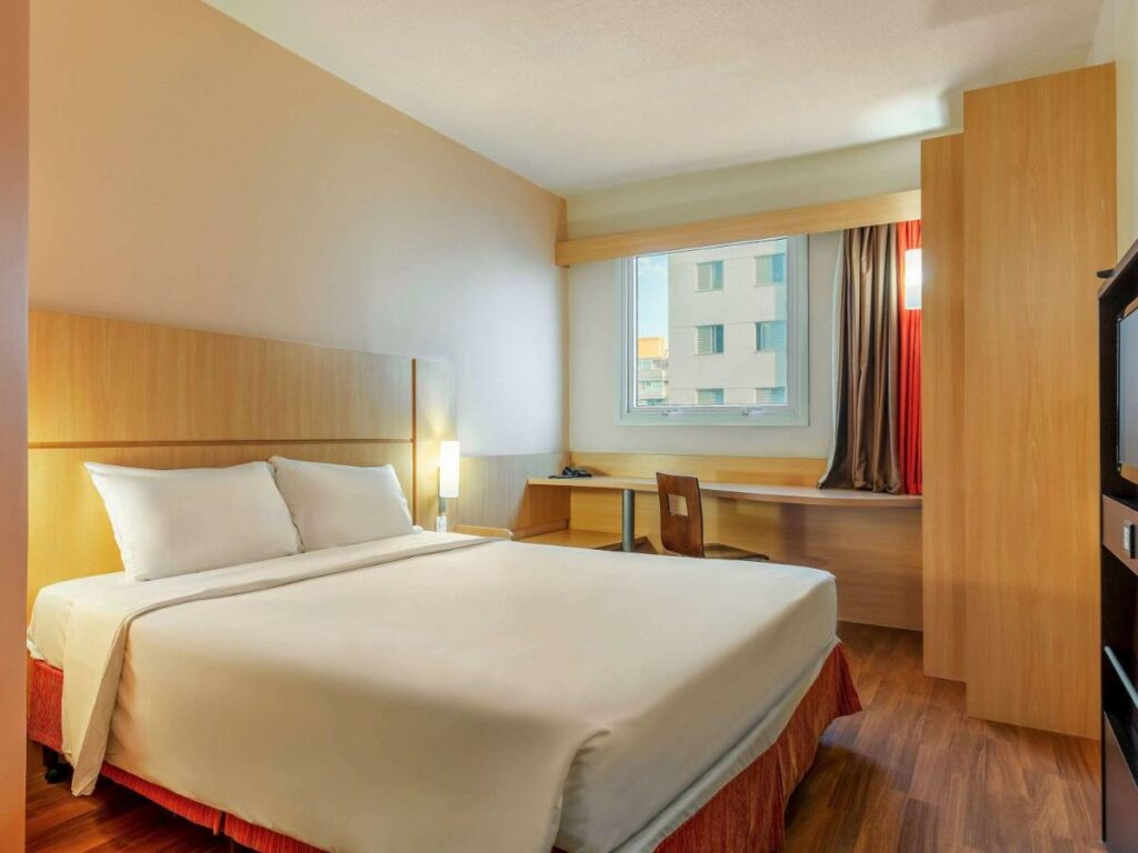 Quarto de hotel com cama de casal com lençóis brancos, mesa de canto em frente à janela. 