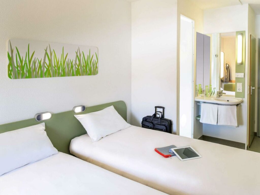 Quarto de hotel com duas camas de solteiro com lençóis brancos, pia do banheiro ao lado e quadro na parede acima das camas. 