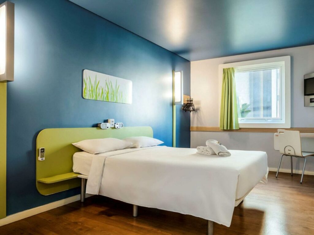Quarto de hotel com cama de casal com lençóis brancos, mesa de canto em frente à janela. 