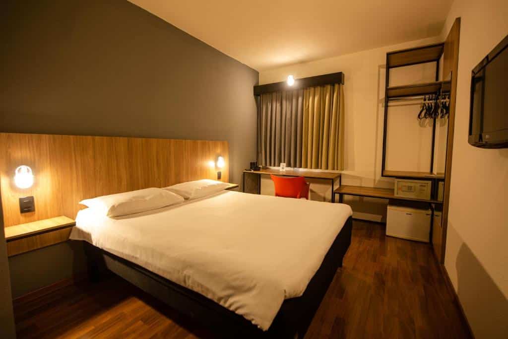Quarto do Ibis Betim, uma alternativa aos hotéis ibis em Belo Horizonte, com cama de casal ampla, TV na parede, armário aberto com cabides, cofre, frigobar, mesa de trabalho com cadeira, e cortina blecaute fechada, estando o quarto iluminado por luzes amarelas