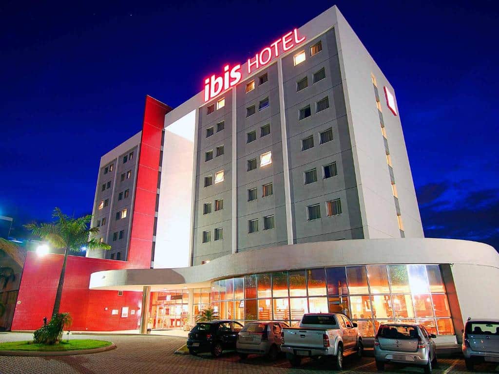 Anoitecer em área externa do Ibis Betim, onde vemos o prédio sob um ângulo baixo, com algumas janelas iluminadas, indicando a presença de hóspedes, e no térreo as paredes são de vidro, com carros parados em frente
