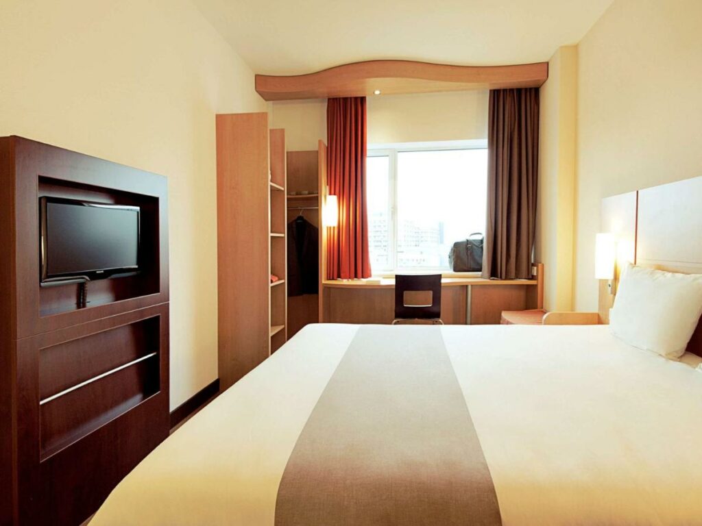 Quarto do ibis Belo Horizonte Liberdade, com TV em painel na parede em frente à cama de casal, armário, mesa de trabalho com cadeira e cortinas blecaute ao fundo