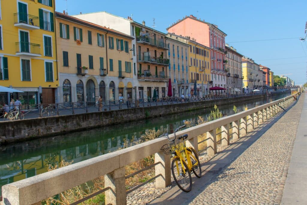 Uma via de pedestres ao lado de um dos canais do bairro de Navigli, onde há um canal e, dos dois lados, há prédios antigos, bicicletas e pessoas andando