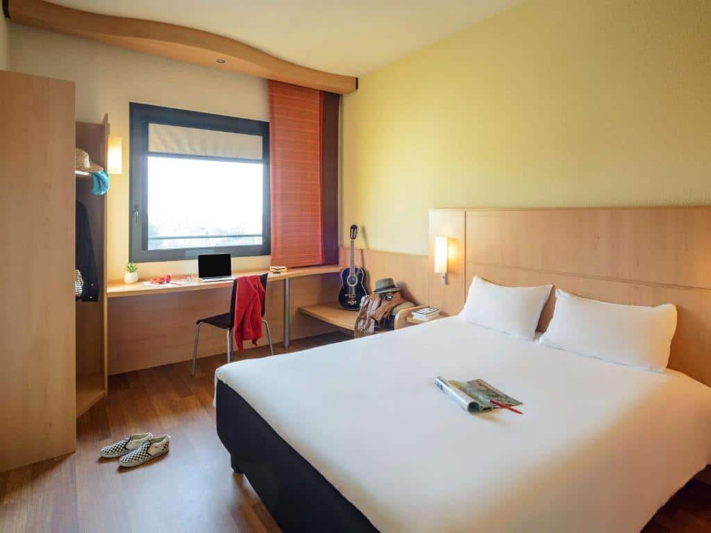 Quarto do Hotel Ibis Milano Malpensa com uma janela ampla, embaixo dela há uma bancada de trabalho e uma cadeira, há um pequeno armário com cabideiro, assim como uma cama de casal, o piso imita madeira e há luminárias presas na cabeceira da cama, para representar hotéis Ibis em Milão