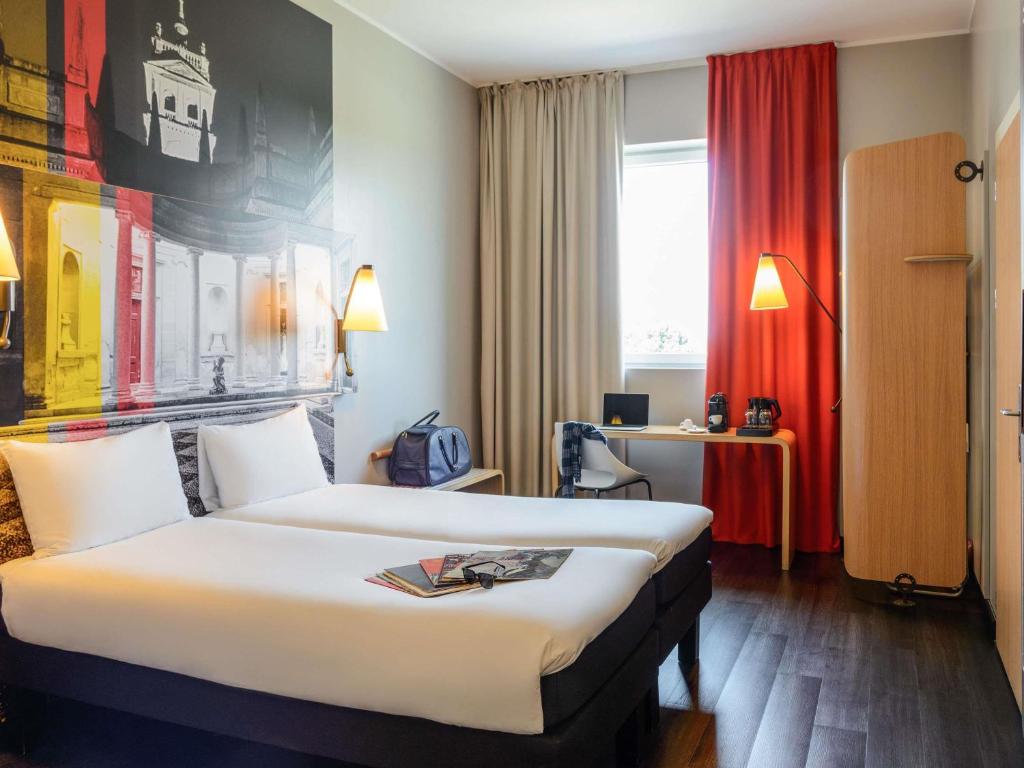 Quarto no ibis Milano Fiera com duas camas de solteiro, uma escrivaninha, uma cadeira, uma janela com cortinas e um mural na parede, para representar hotéis ibis em Milão.