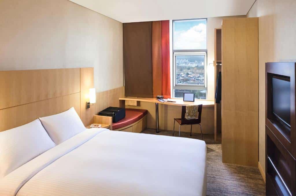 Quarto do Hotel Ibis Milano Ca' Granda com uma cama de casal, uma mesa de escritório e uma janela logo acima dela, há um armário com cabideiro e uma televisão em frente a cama, para representar hotéis Ibis em Milão