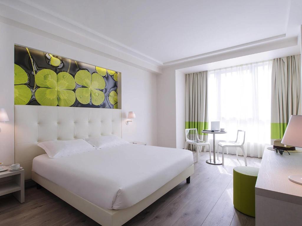 Quarto no ibis Styles Milano Est Settala com uma cama de casal, uma janela grande, uma parede com arte de nenúfares e uma pequena mesa com duas cadeiras, para representar hotéis ibis em Milão.