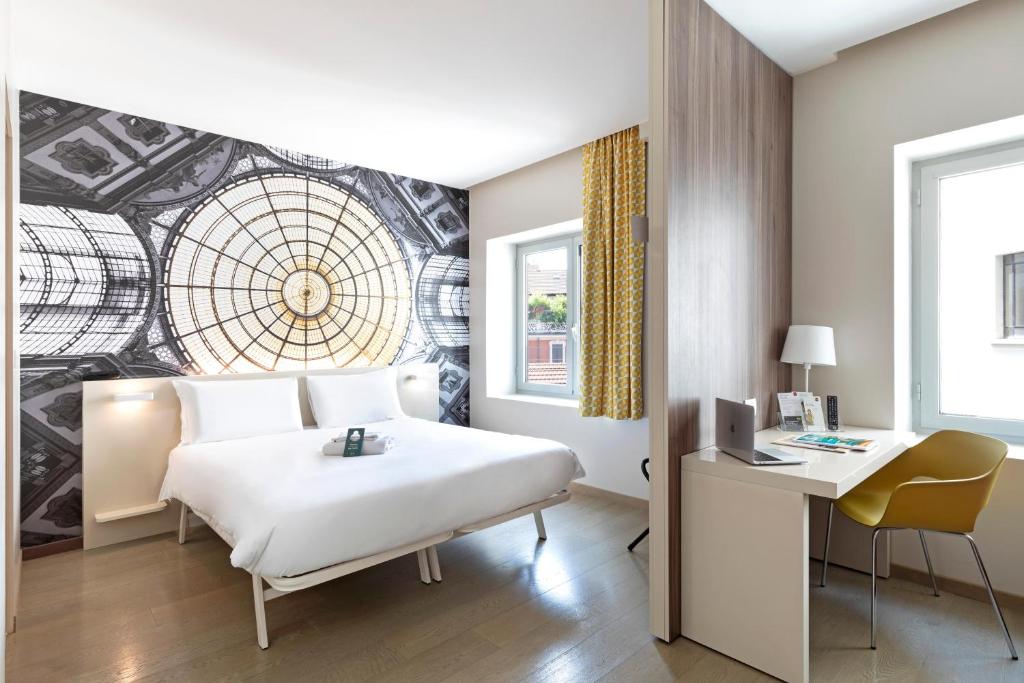 Quarto no B&B Hotel Milano Central Station com uma cama de casal, arte de parede grande, escrivaninha com cadeira, luminária e duas janelas que permitem a entrada de luz natural, para representar hotéis ibis em Milão.
