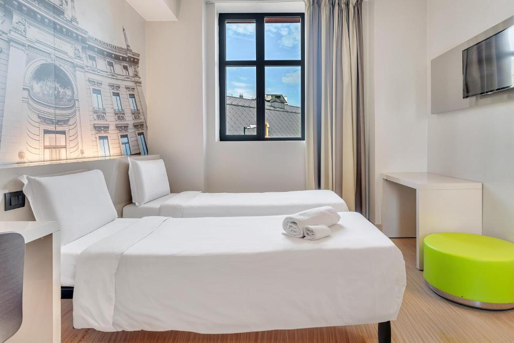 Quarto no B&B Hotel Milano San Siro com duas camas de solteiro, um mural na parede, uma janela, uma TV de tela plana e um banco redondo verde, para rperesentar hotéis ibis em Milão.