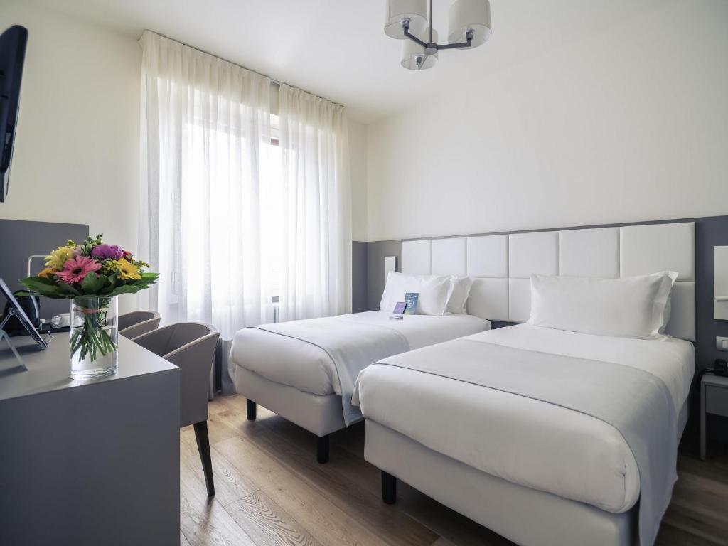 Quarto no Hotel Mercure Milano Solari com duas camas de solteiro, roupa de cama branca, uma escrivaninha com um vaso de flores, duas cadeiras e uma janela com cortinas transparentes, para representar hotéis ibis em Milão.