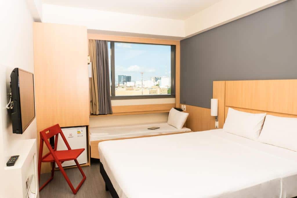 Quarto do ibis Rio Porto Atlantico com uma cama de casal e uma de solteiro com lençóis brancos, janela de vidro, cadeira marrom, frigobar e uma tv, ilustrando o post hotéis Ibis no Rio de Janeiro.