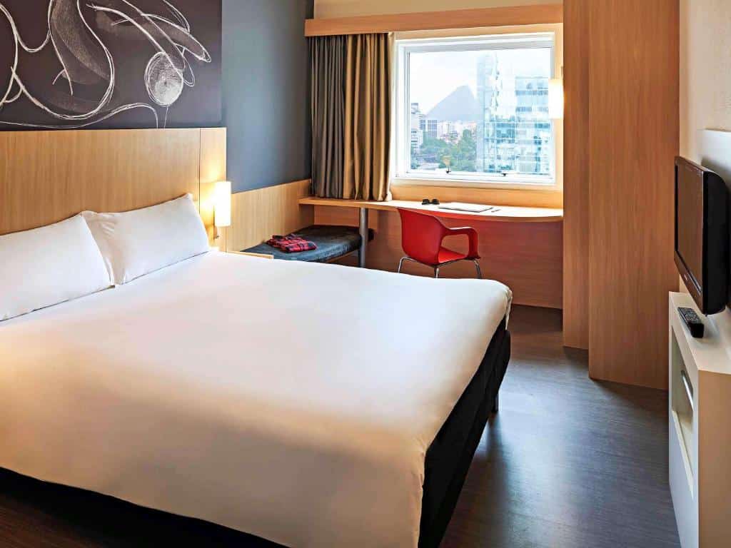 Quarto do ibis Rio de Janeiro Centro com uma cama de casal branca, detalhes brancos na parede preta, escrivaninha com uma cadeira vermelha, janela de vidro e uma tv na parede, ilustrando post hotéis Ibis no Rio de Janeiro.
