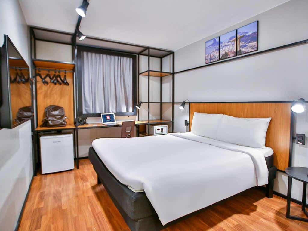 Quarto do hotel ibis Copacabana Posto 2 com cama de casal branca, chão de madeira, escrivaninha, frigobar. cabides e uma tv, ilustrando post hotéis Ibis no Rio de Janeiro. 