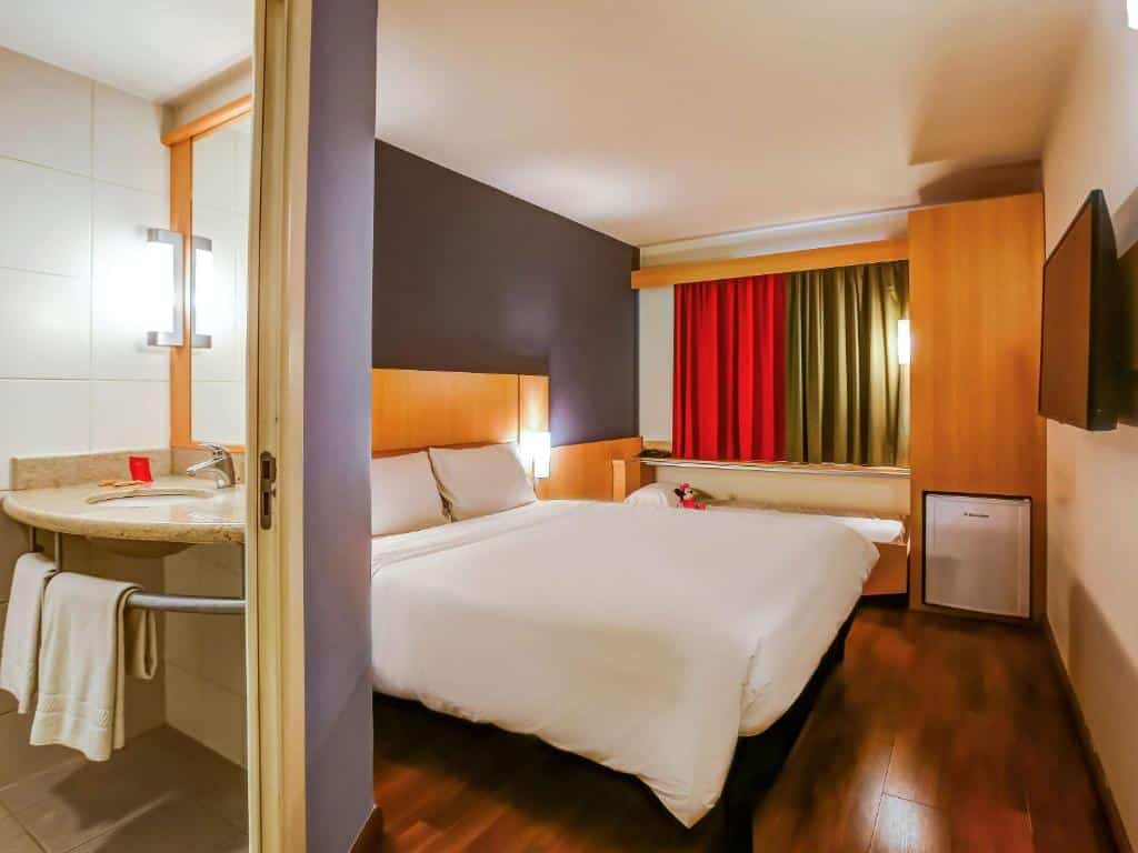 Quarto do ibis Rio de Janeiro Botafogo com uma cama de casal branca e uma cama de criança, cortina colorida, frigobar, tv e na imagem aparece parte da pia do banheiro, ilustrando post hotéis Ibis no Rio de Janeiro.