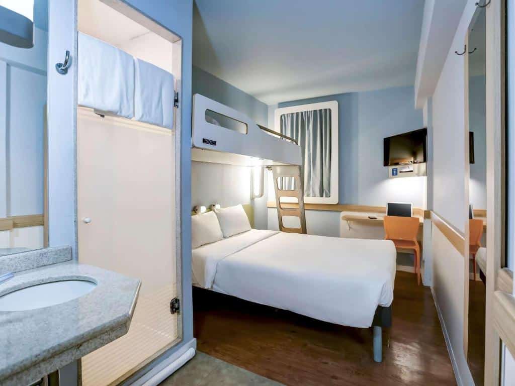 Quarto do ibis budget RJ Copacabana com uma cama de casal e em cima um beliche de solteiro, uma escrivaninha com cadeira amarela, uma tv e parte da pia do banheiro, ilustrando post hotéis Ibis no Rio de Janeiro.