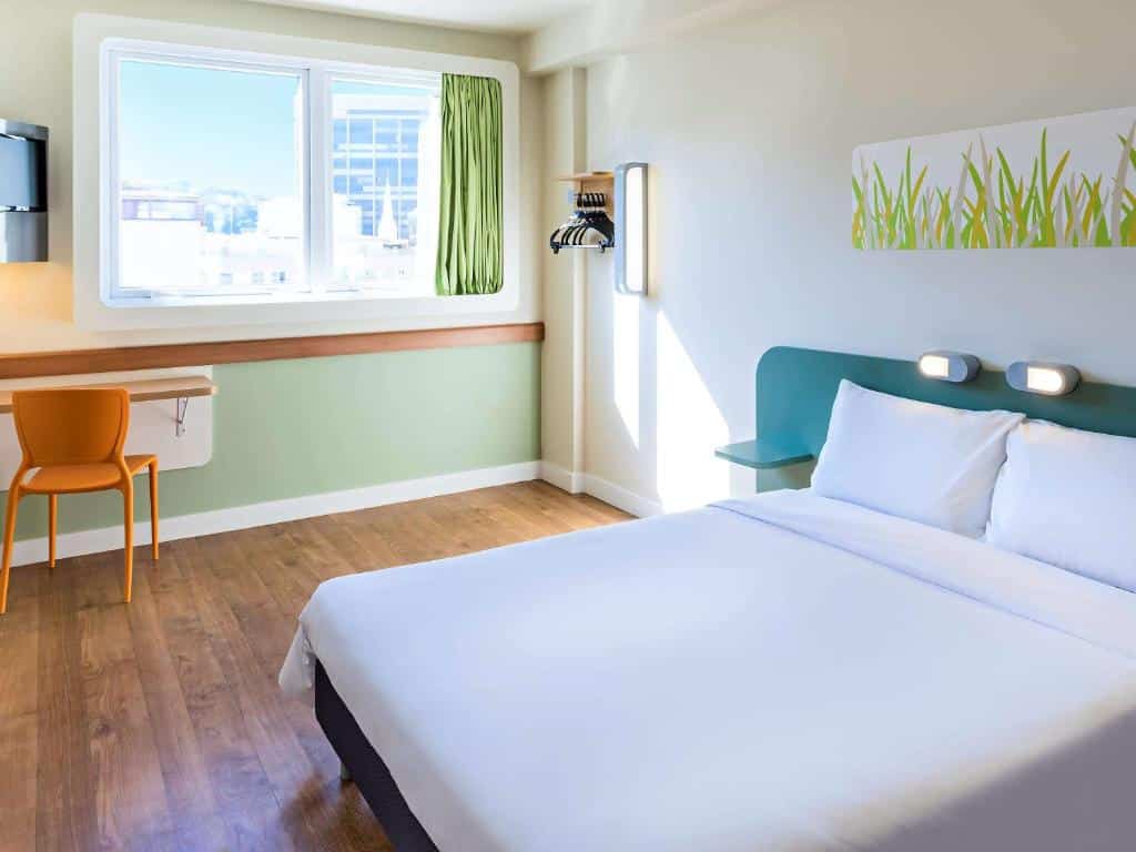 Quarto do ibis budget RJ Praia de Botafogo com cama de casal branca, chão de madeira, escrivaninha com cadeira amarela, tv, janela de vidro e detalhes de grama verde na parede, ilustrando post hotéis Ibis no Rio de Janeiro.