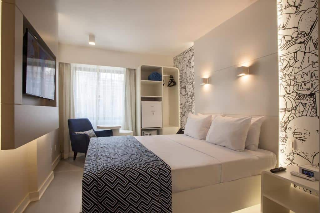 Quarto do ibis Styles Rio de Janeiro Botafogo com uma cama de casal, tv na parede, cadeira azul, janela com cortina branca, frigobar e paredes brancas com detalhes pretos, ilustrando post hotéis Ibis no Rio de Janeiro.