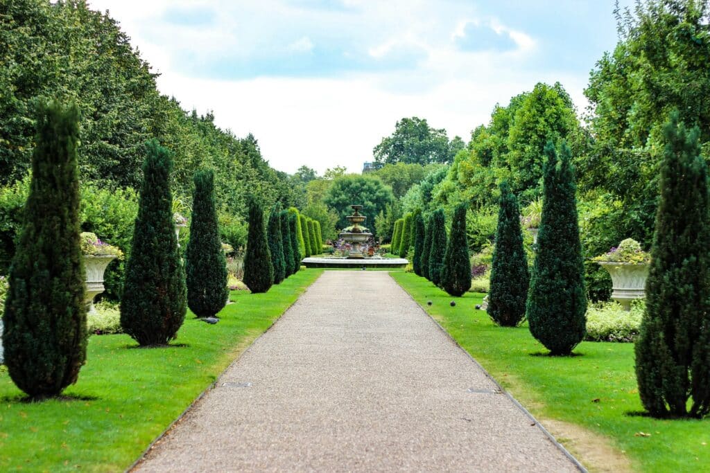 Vista da parte interna do Regents Park com muitas árvores desenhadas, um corredor de concreto que leva até uma fonte, com tudo cercado com gramado, flores e árvores