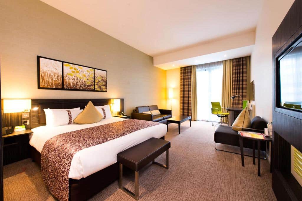 Quarto do Holiday Inn London - Whitechapel, an IHG Hotel com uma cama de casal, um sofá, uma janela com cortinas, uma poltrona, uma televisão e uma mesa de escritório com uma cadeira, para representar hotéis Mercure em Londres