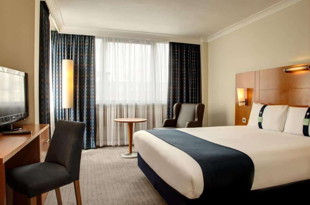 Quarto do Holiday Inn London Bloomsbury, an IHG Hotel com uma cama de casal, uma janela com cortinas, uma poltrona, uma mesa de escritório com cadeira e uma televisão