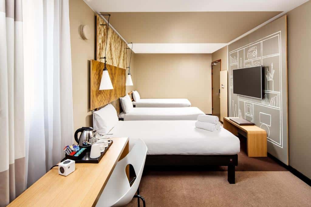 Quarto do ibis London City - Shoreditch com três camas de solteiro, carpete marrom claro, uma televisão, uma mesa de escritório com alguns itens de café sob ela e uma cadeira, para representar hotéis Mercure em Londres