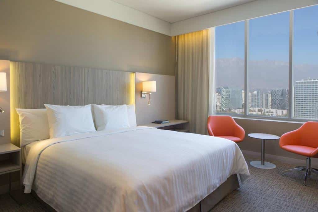 Quarto do Courtyard by Marriott Santiago Las Condes com cama de casal do lado direito duas cômodas ao lado da cama e duas cadeiras laranjas e ao fundo janela, para representar hotéis Mercure em Santiago.