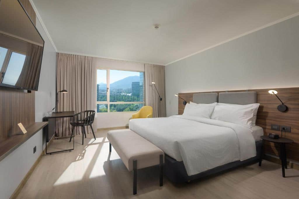 Quarto do Novotel Santiago Las Condes com cama de casal do lado direito com uma mesa do lado direito da cama, uma poltrona ao pé da cama e uma TV em frente a cama, para representar hotéis Mercure em Santiago.