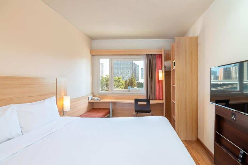 Quarto do ibis Santiago Estacion Central com cama de casal do lado esquerdo, com uma cômoda em frente a cama com TV em cima. Do lado esquerdo mesa de trabalho com janela, para representar hotéis Mercure em Santiago.