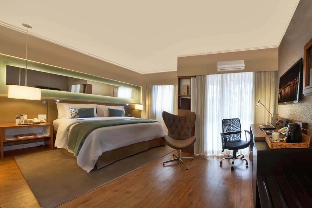 Suíte no hotel Clarion Faria Lima. O ambiente é moderno e aconchegante. A cama é larga e possui lençóis brancos, travesseiros brancos e alguns cinzas e uma manta verde militar sobre o pé da cama. No chão, há um tapete fino e marrom claro. Do lado direito da cama é possível ver uma mesa de cabeceira com alguns livros, telefone e outros itens. Um abajur está do lado direito da cama, enquanto uma luminária pendurada está no lado esquerdo. Na parede, há um espelho longo que percorre toda a área na vertical. No lado esquerdo, há janelas com cortinas claras, e um ar-condicionado em cima. De frente para a cama, encostada na parede, tem uma escrivaninha marrom com um notebook, um abajur e uma bandeja para café. Uma cadeira de escritório preta está próxima ao móvel e outra cadeira marrom também está do lado. Há uma tv na mesma parede da escrivaninha. Foto para representar o post sobre hotéis na Faria Lima em SP.