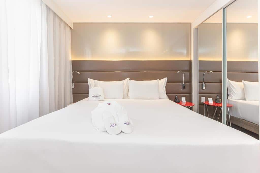 Foto do quarto do hotel Mercure. A cama, travesseiros e cortina são brancos. Há uma roupa de banho e pantufas do hotel disponíveis na cama. Uma mesa de cabeceira pequena na cor vermelha está do lado direito da cama. Ainda no lado direito, podemos ver um guarda-roupa espelhado. Foto para representar o post sobre hotéis na Faria Lima em SP.
