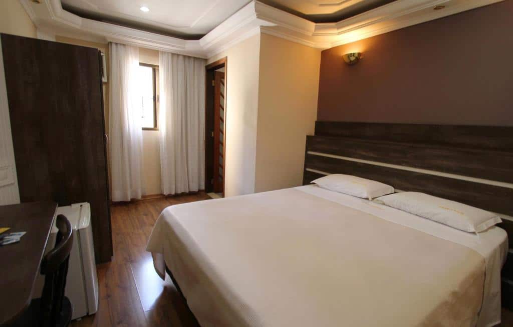 Foto do quarto do Gran Chevalier Hotel. Podemos ver a cama com lençóis e travesseiros brancos, e uma cabeceira de madeira escura. A parede atrás está pintada na cor marrom. De frente para a cama, é possível ver uma mesa e cadeira também na cor marrom, e um frigobar do lado. Do lado da mesa, tem um guarda-roupa. também, marrom. A janela tem cortinas brancas. Foto para ilustrar o post sobre hotéis na Faria Lima em SP.