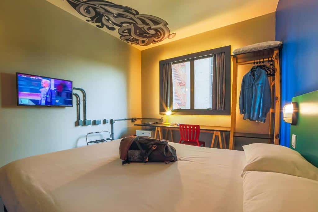 Quarto do hotel ibis Styles Faria Lima. A cama é de casal, com lençóis e travesseiros brancos. Uma mochila preta está em cima da cama. A parede atrás é colorida em azul, verde e vermelho. Do lado, há uma arara de roupa de madeira, uma camisa azul e alguns cabides estão pendurados. Do lado, uma escrivaninha de madeira, com um abajur e uma cadeira vermelha estão disponíveis para uso. De frente para a escrivaninha, está a janela do quarto com cortinas pretas. Uma tv de tela plana está de frente para a cama, e nela passa algum jogo. O teto também está decorado com uma arte moderna em cores pretas e cinzas. Imagem para representar o post sobre hotéis na Faria Lima em SP.