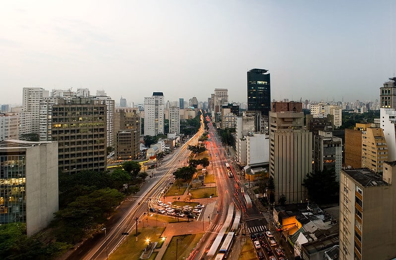 Imagem da Avenida Brigadeiro Faria Lima em São Paulo. O céu está nublado, e é quase noite. A avenida está no centro da foto, muitos carros passam e os faróis iluminam a cidade. Dos lados, vemos inúmeros edifícios, hotéis e empresas.