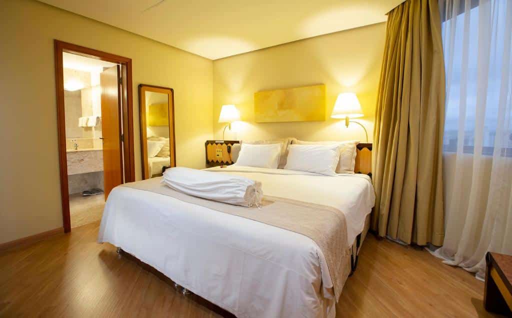 Foto do quarto do hotel Golden Tower na Faria Lima. Uma cama grande e com lençóis brancos está no centro da foto. Há abajures fixos na parede, um em cada lado da cama, e um quadro amarelo no centro. Na direita, tem uma janela com uma cortina parte bege e outra parte branca. Na esquerda, há um espelho do lado da cama e uma porta para o banheiro. Foto pra representar o post sobre hotéis na Faria Lima em SP.