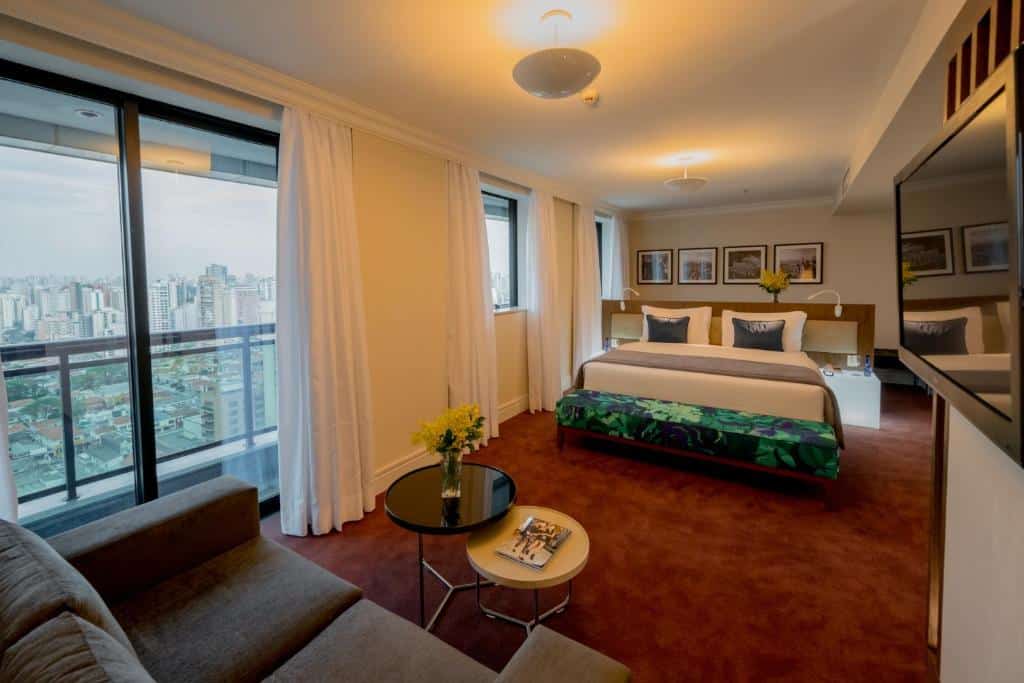Foto do quarto do Blue Tree Premium Faria Lima. Perto da câmera, está o sofá marrom escuro e de frente duas mesas de centro pequenas que estão juntas uma da outra. Do lado esquerdo, tem uma janela com bordas pretas e cortinas pretas. Do lado direito, tem uma tv fixa na parede. O chão do quarto tem um carpete vermelho. No outro lado do quarto, tem uma cama grande com lençóis e travesseiros brancos, e outros dois travesseiros mais escuros. Uma manta cinza está sobre a cama. Um pequeno sofá-cama está em frente da cama, estampado de folhas tropicais. quadro quadros decoram a parede. Foto para representar o post sobre hotéis na Faria Lima em SP.