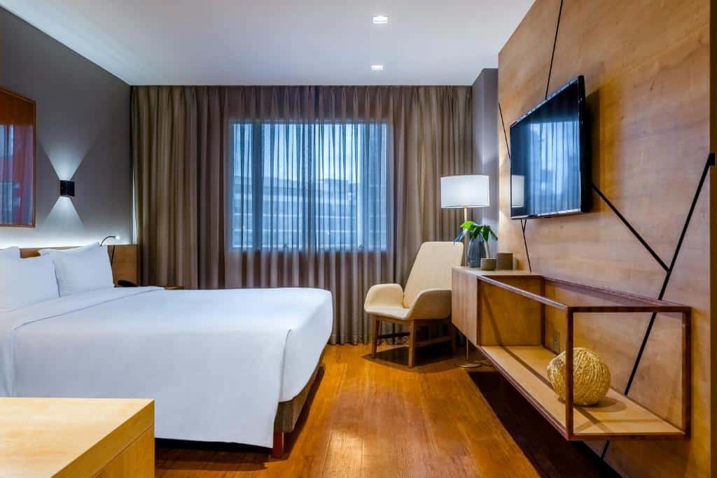 Foto da suíte para representar o post sobre hotéis na Faria Lima em SP. A cama possui lençóis e travesseiros brancos. O piso do quarto é uma madeira marrom clara, e um painel do mesmo padrão decora a parede de frente pra cama,. Uma TV e uma mesa moderna estão de frente para a cama. Uma poltrona bege está do lado de um abajur do lado da janela, que possui cortinas cinzas. 