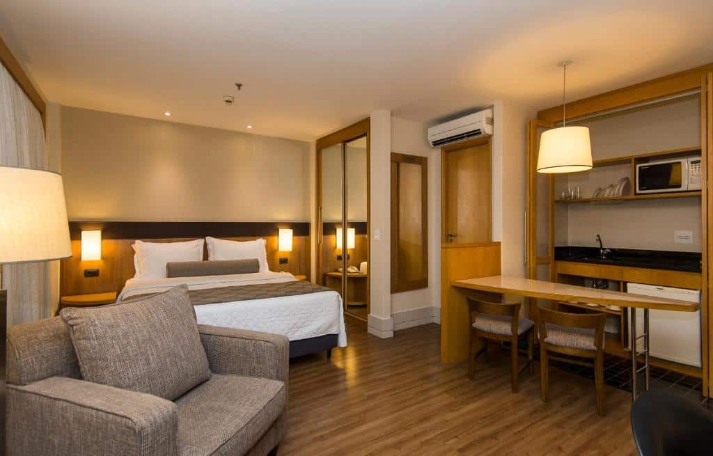 Imagem do quarto do hotel Transamerica Executive. Vemos uma poltrona cinza e um abajur do lado. Atráas, está a cama de casal com lençóis e travesseiro brancos, e uma manta marrom está em cima dela. Dois abajures estão fixos na parede em ambos os lados da cama. Do lado direito, tem um guarda-roupa com portas espelhadas. Do lado direito, uma janela com cortinas brancas. Em outro canto do quarto, há uma mini cozinha equipada com frigobar, pia, micro-ondas e alguns prato. Uma pequena mesa está no ambiente com duas cadeiras e uma luminária está em cima da mesma. O ar condicionado do quarto está em cima da porta de entrada do quarto, e tem um espelho do lado. Foto para ilustrar o post sobre hotéis na Faria Lima em SP.
