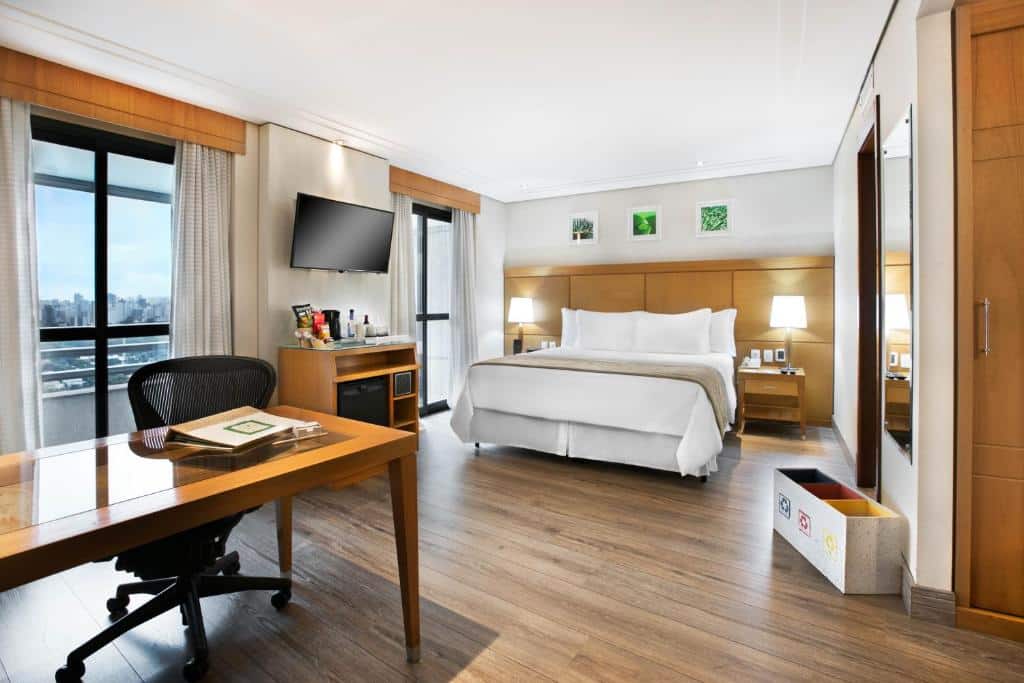 Foto do quarto do hotel Meliá Jardim Europa. A suíte é espaçosa, a mesa é grande e seus lençóis e travesseiros são brancos. Uma manta bege está em cima da cama. Há dois abajures em cada lado da cama sobre uma mesa de cabeceira. Três quadros com artes verdes decoram a parede. Do lado esquerdo, tem as janelas do hotel, com bordas pretas e cortinas claras. Entre essas duas janelas, há uma mesa comportando alguns itens do quarto e uma TV em cima, fixa na parede. Uma mesa de madeira está disponível no quarto, com uma cadeira preta giratória acompanhando. Há um espelho no quarto e alguns lixos de reciclagem embaixo. Foto para representar o post sobre hotéis na Faria Lima SP.