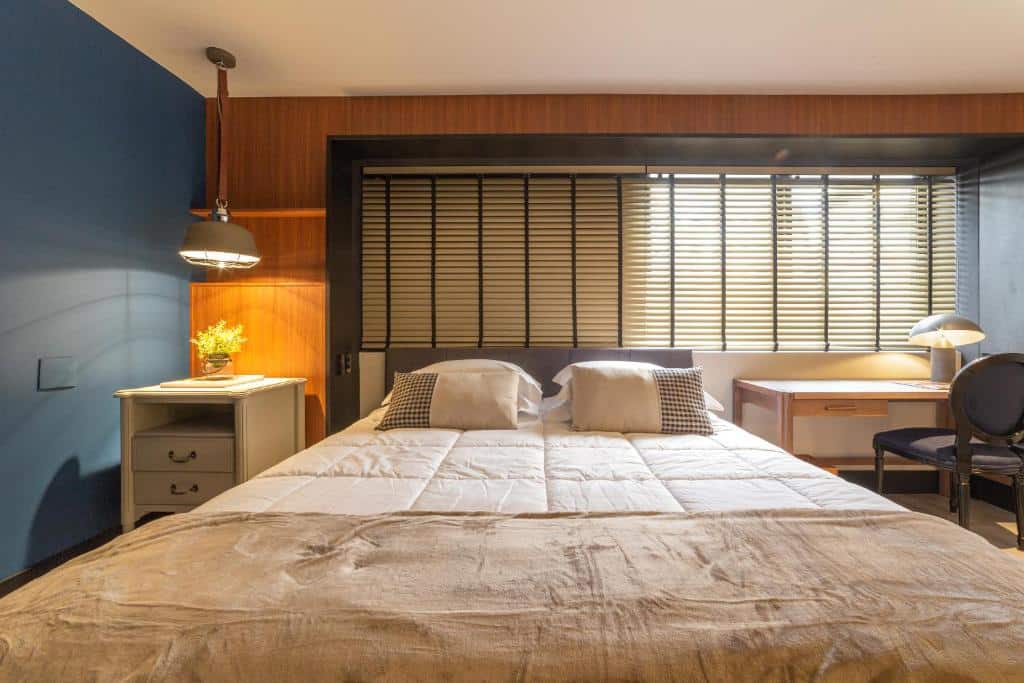 Foto do quarto no Cyclinn Faria Lima Itaim. A cama é de casal, uma manta bege está sobre ela. Quatro travesseiros acompanham o móvel. Do lado esquerdo da cama, há uma mesa de cabeceira de madeira clara com duas gavetas. Há uma plantinha em cima e um abajur pendurado em direção à ela. A parede do lado esquerdo é azul marinho. Do lado direito da cama, há uma pequena escrivaninha bege com uma cadeira azul, e um abajur está em cima dela. De frente, há uma janela com persianas. Foto para ilustrar o post sobre hotéis na Vila Olímpia SP.